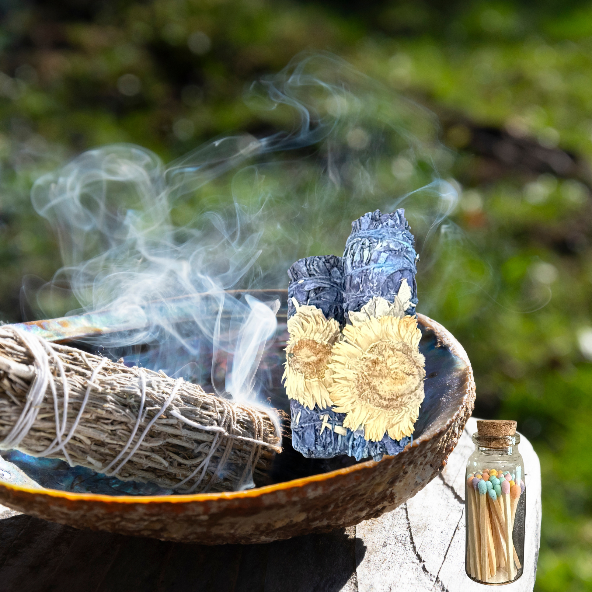 Incense sticks with smoke, a blue container with sunflower design, and a bottle of matches on a natural background.