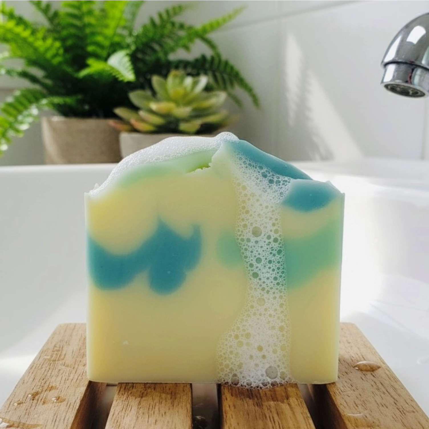 Bar of soap with green and blue swirls on a wooden block, with a bathtub and plants in the background.
