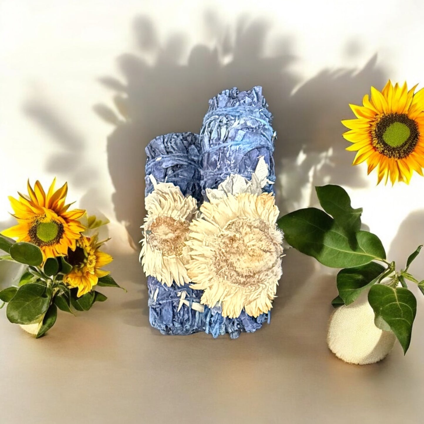 A breakable blue-colored white sage smudge stick decorated with artificial flowers, displayed alongside fresh sunflowers and greenery against a grey background.