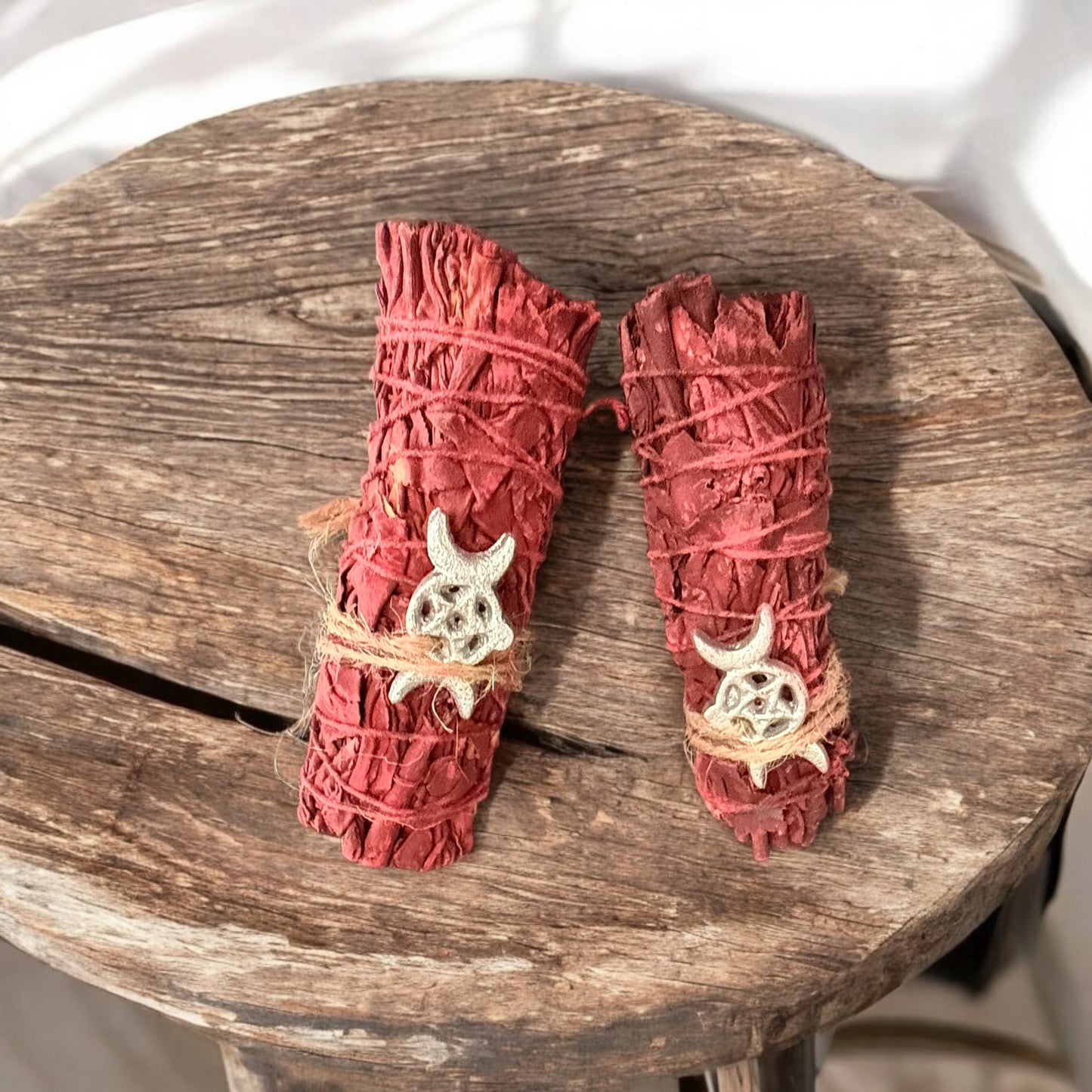 Two Dragon's Blood Sage smudge sticks with tied strings and adorned with a silver Triple Moon charm, placed on a wooden surface.