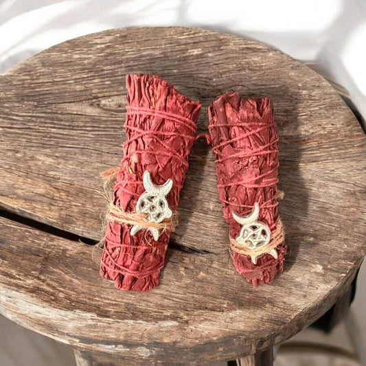 Two Dragon's Blood Sage smudge sticks with tied strings and adorned with a silver Triple Moon charm, placed on a wooden surface.