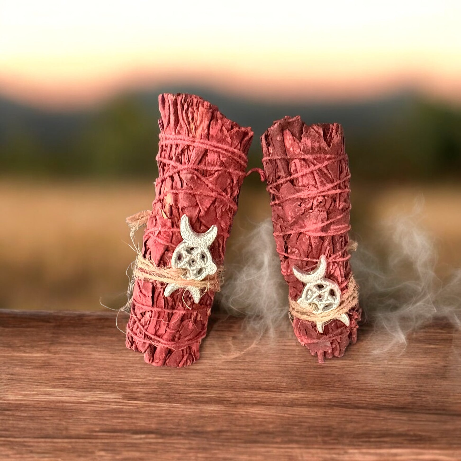 Two bundles of red sage tied with string, featuring a moon crest design, on a wooden surface with smoke rising from the ends.