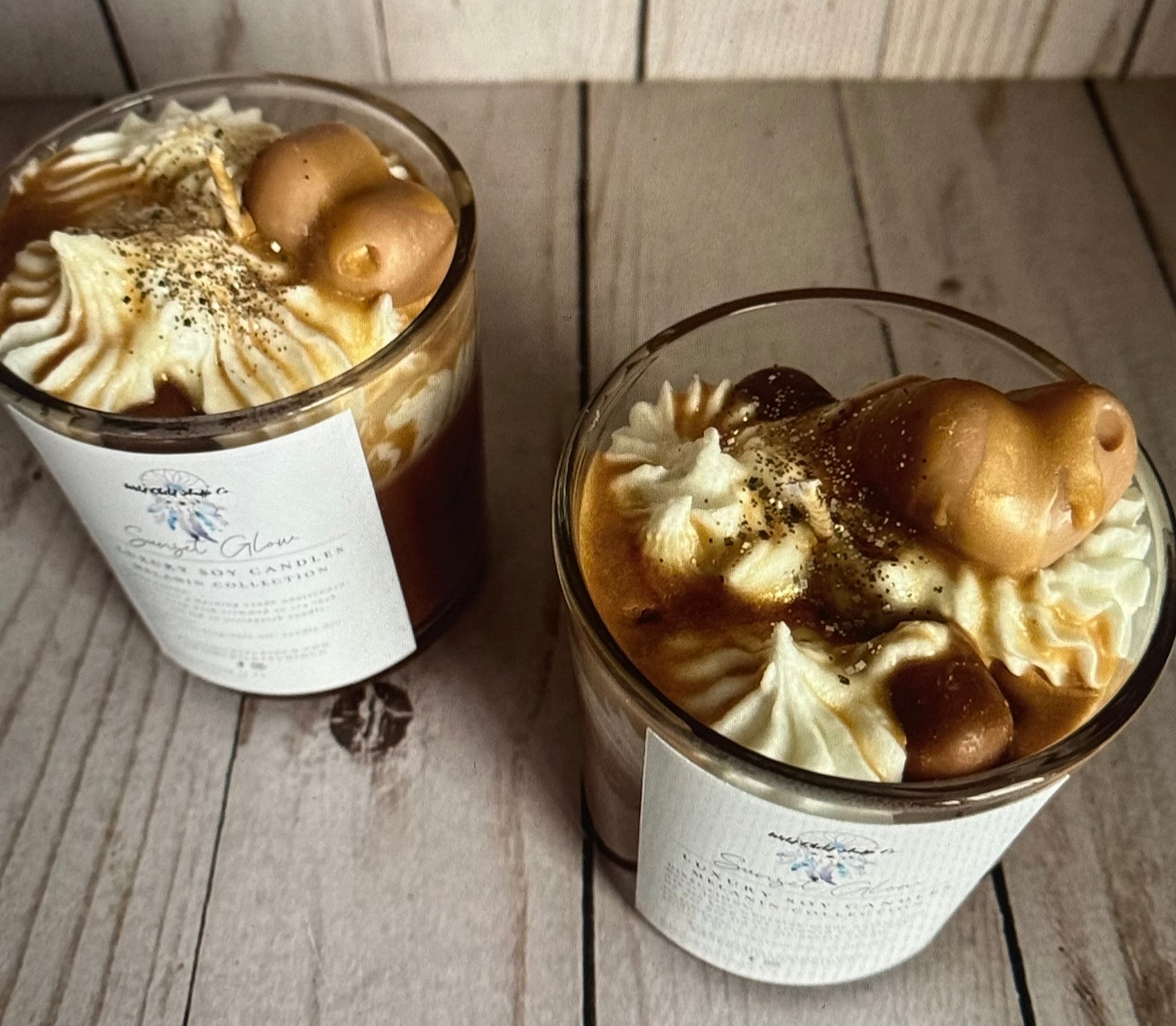 Two candles with medium brown hue and white wax, adorned with heart wax embeds, gold drizzle, and glitter, in clear glass jars with bamboo lids, on a wooden surface.
